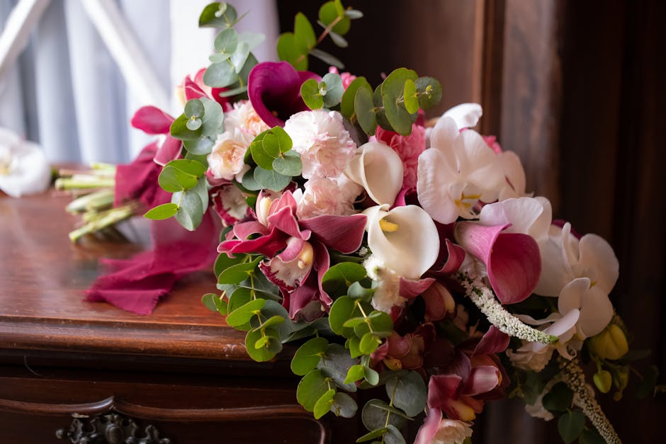 A floral bouquet featuring creamy white and deep magenta calla lilies, soft pink and white roses, and delicate white orchids, accented with lush green eucalyptus leaves. The arrangement is resting on 