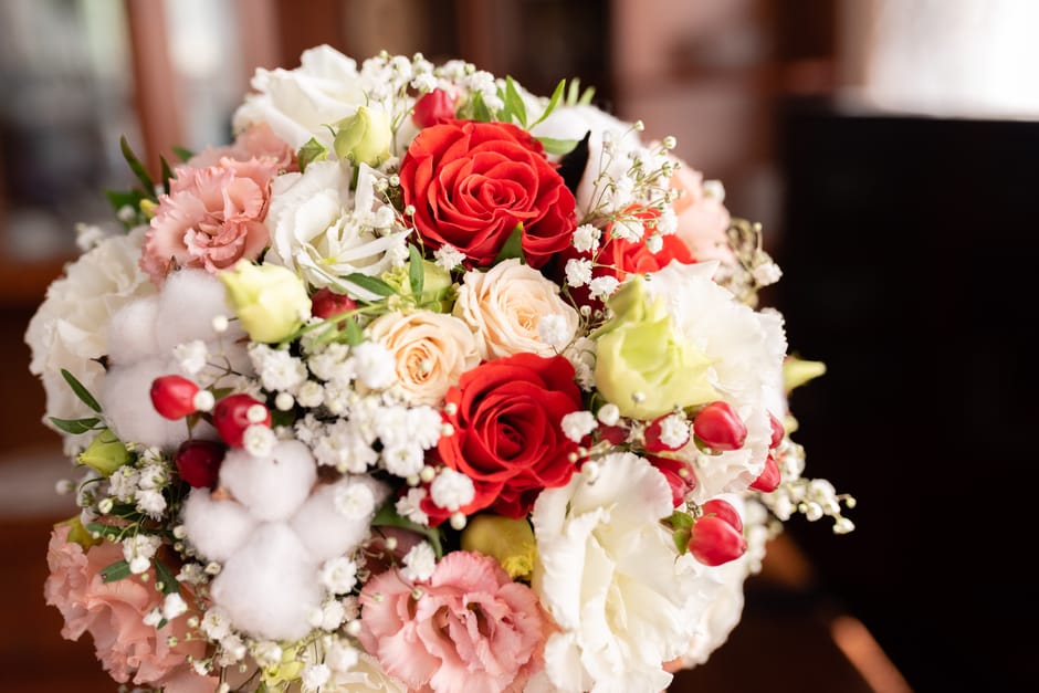 A close-up of a floral bouquet arranged with fresh red roses, pale peach roses, white lisianthus, pink and white carnations, sprigs of baby's breath, and cotton branches, all elegantly displayed with 
