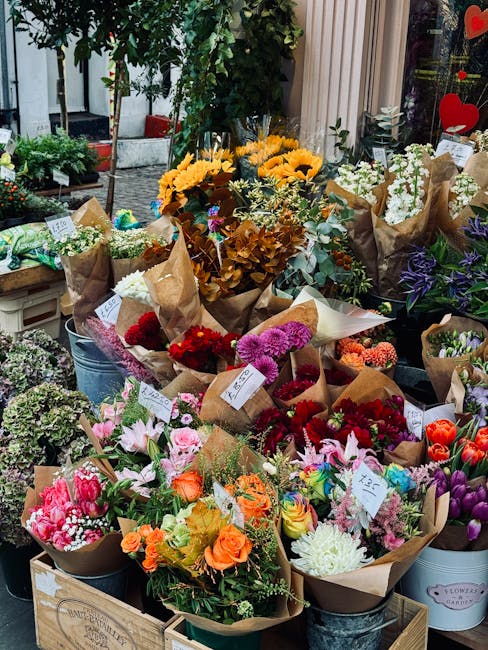 A vibrant display of fresh flower bouquets arranged outdoors on a shop's table. The arrangements include sunflowers with bright yellow petals, pink lilies with delicate petals, red and purple chrysant