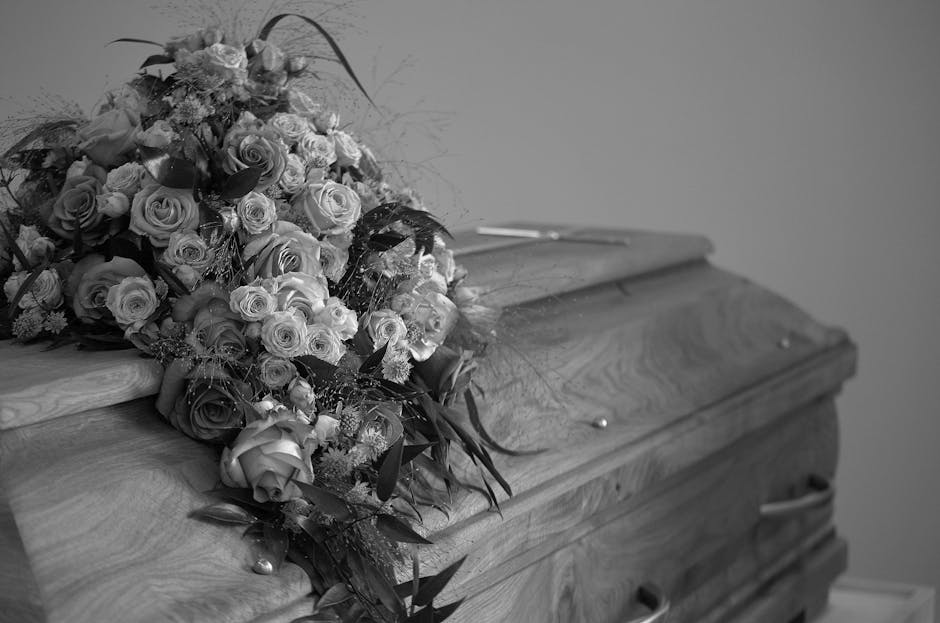 A large funeral floral arrangement resting on a wooden coffin, consisting of a wide array of fresh roses in shades of white, cream, and pale pink, interspersed with dark green foliage and delicate bab