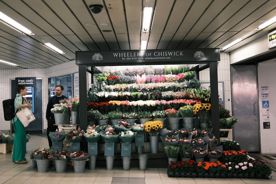 A vibrant flower display at Wheeler of Chiswick flower stall inside a subway station, featuring a variety of fresh flowers arranged in buckets and wrapped bouquets. The arrangement includes white, pin