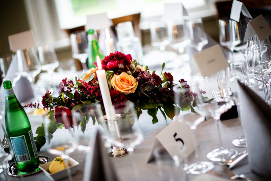 A floral arrangement featuring a bouquet of fresh roses in soft peach and deep burgundy, complemented by smaller purple and red blossoms, arranged in a silver vase on a table set for a formal event. T