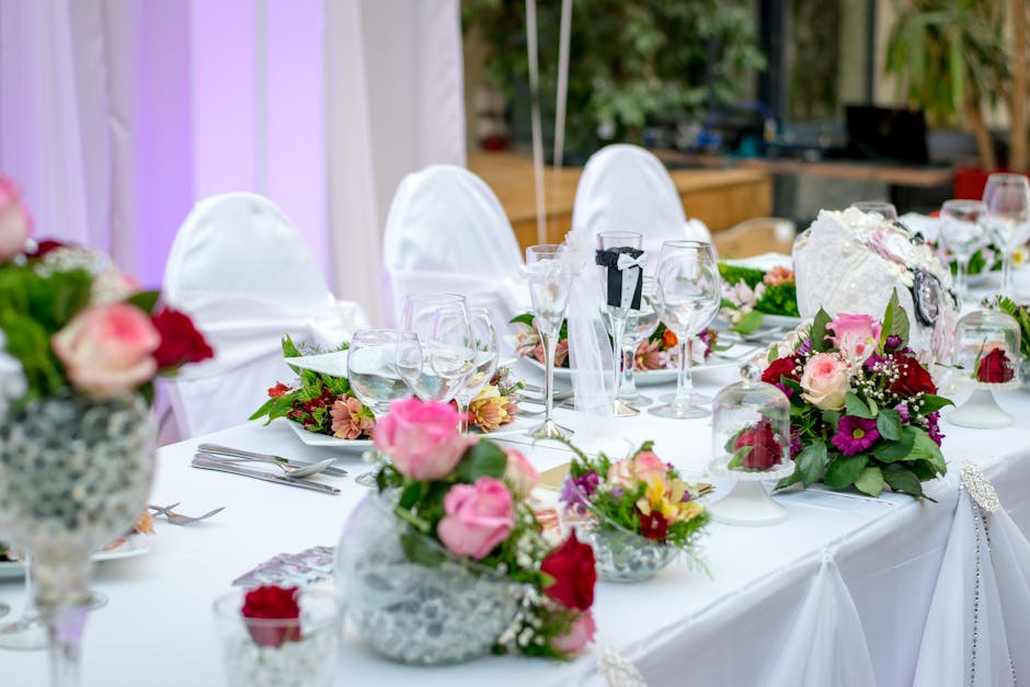 A beautifully arranged banquet table set for a wedding event, featuring multiple floral arrangements with pink, red, and purple flowers, including roses, anemones, and baby's breath, placed in elegant
