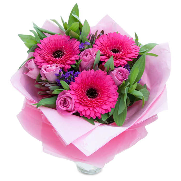 I see this bouquet as a small, perfumed tableau of pinks assembled with intention and warmth. At its visual heart are three fuchsia Gerbera daisies, bold and open, each petal catching the light and pointing outward from a strikingly dark centre that lends depth. Encircling those confident forms are roses in a muted lavender-pink, their petals layered in soft, almost dusty spirals that suggest tenderness and quiet affection. Between the larger flowers, clusters of purple statice drift like little fields of wildflower color, bringing playful texture and a cool counterpoint to the dominant pinks. The greens are generous and grounding - broad salal-like leaves and eucalyptus tones that read as fresh-cut from a nearby garden, their matte sheen and faint herbal scent anchoring the composition. The bouquet is wrapped in pale pink tissue with an outer leaf of translucent, slightly crinkled fuchsia-pink cellophane that shimmers under gentle light, folded with careful, artisanal hands. The clear glass vase rim is visible at the bottom, indicating the piece is ready to stand on a kitchen table or shelf, perhaps to greet a guest after a morning walk through Holland Park or a browse at Portobello Road market. This arrangement evokes celebration in a soft key - birthdays, a thoughtful anniversary, or a spontaneous 'thinking of you' - and carries the quiet assurance of quality that comes from a florist who knows the neighbourhood and respects the rhythm of seasonal flowers.